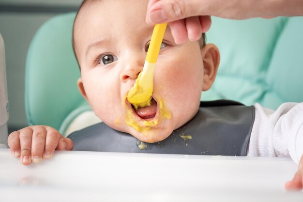 Mom feeds the baby with a spoon of vegetable puree at the children's feeding table Baby's appetite healthy nutrition introduction of complementary foods Copyspace mock up