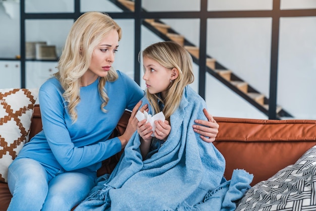 Mom helping her little daughter with her ill on sofa at home