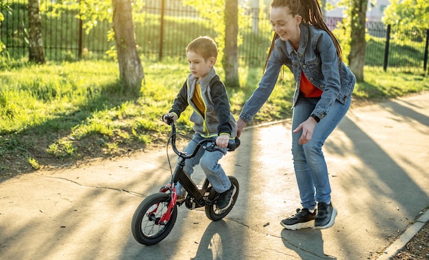 Mom helps a child boy learn to ride a twowheeled bicycle in the park A pleasant children's summer sports vacation