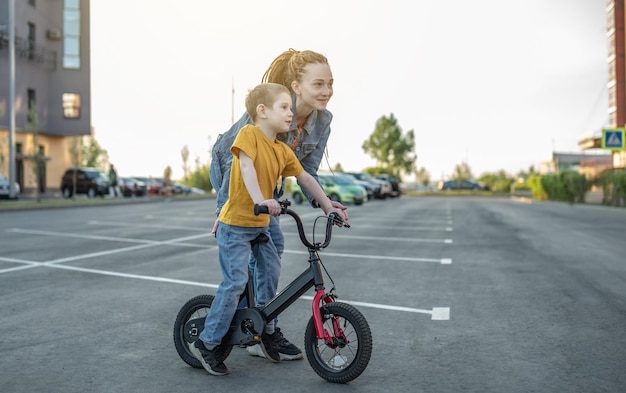 Mom helps a child boy learn to ride a twowheeled bicycle in the park A pleasant children's summer sports vacation