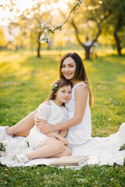 Mom hugs her daughter sitting on a blanket in a spring blooming garden Maternal love for a child