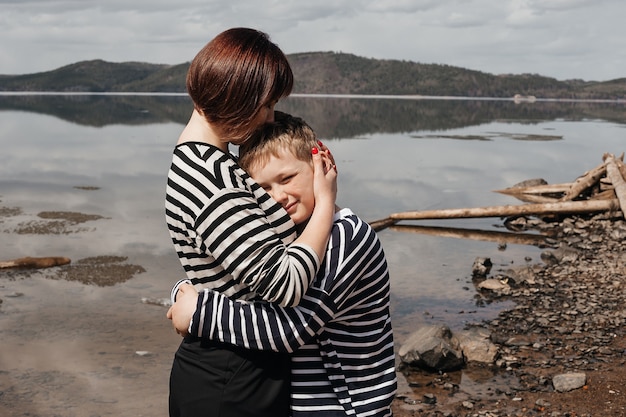 Mom hugs her son on the riverbank. A beautiful, modern mother with a happy son in striped vests.