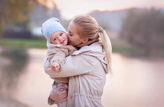 Mom Hugs and Kisses smiling Baby at sunset on nature lake. Blurred background, copy space, beige color. Mother loves her son