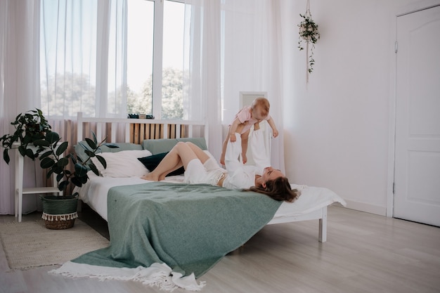 Mom is lying on the bed and playing with her baby daughter in a pink bodysuit