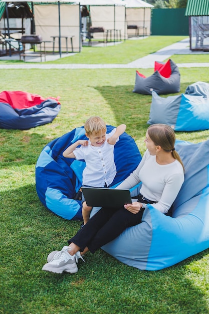 Mom is on maternity leave Charming young woman with little son working outdoors in summer park with laptop while sitting on chair on green grass