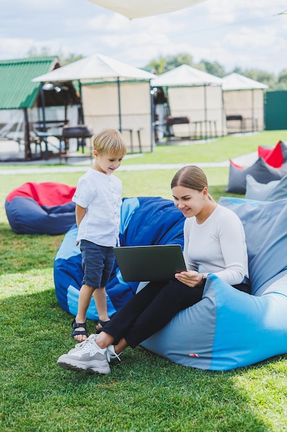 Mom is on maternity leave Charming young woman with little son working outdoors in summer park with laptop while sitting on chair on green grass