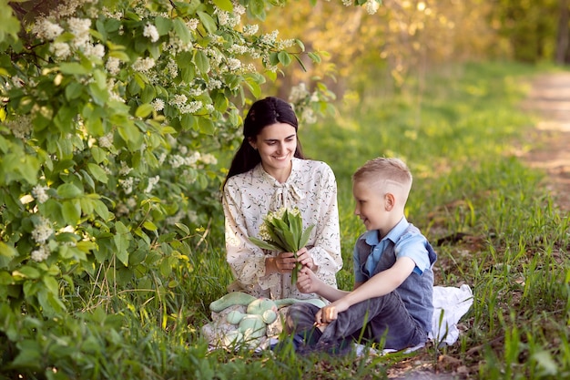 Mom is sitting next to her son on the grass the boy is holding flowers in his hands