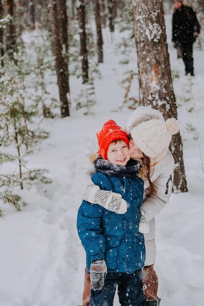 Mom kisses her son in the winter forest. Mom walks with her son in a snowfall in the forest. Happy winter holidays with snow. Winter walks in the forest. Mom's love for her son.