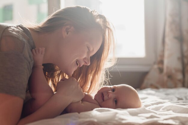 Mom kisses and hugs a smiling newborn baby lying on the bed young woman with a child at home