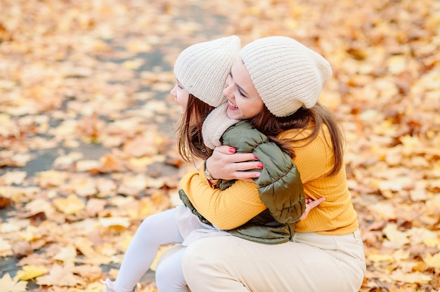 Mom in a knitted hat gently looks at her daughter of five years old holding her in her arms in the fall against the background of fallen yellow leaves