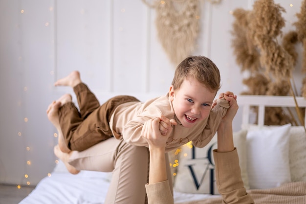 Mom and a little boy play cuddle and make a plane at home on a bed in natural brown tones a happy family with a small child has fun together