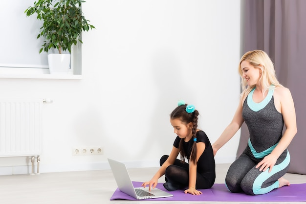 Mom and little daughter are doing gymnastics on the mat at home. They do yoga. They are fun because they have a happy family. Poses they are looking at the laptop.