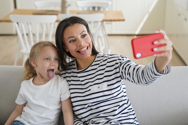 Photo mom little daughter take selfie photo by phone show tongues together having fun sitting on sofa