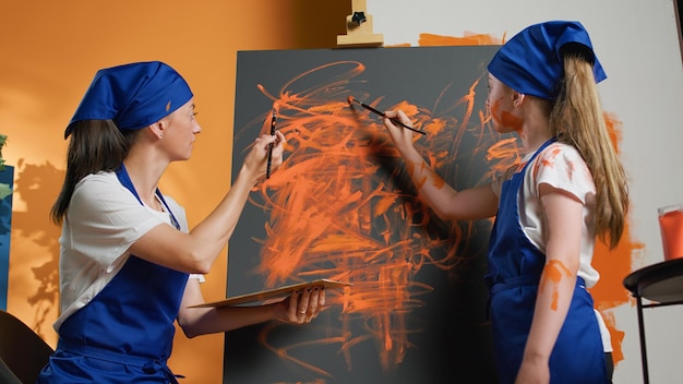 Mom and little girl painting artwork on canvas with aquarelle palette and paintbrush. Young people learning to paint design with watercolor dye on mixing tray, using artistic vision and skills.