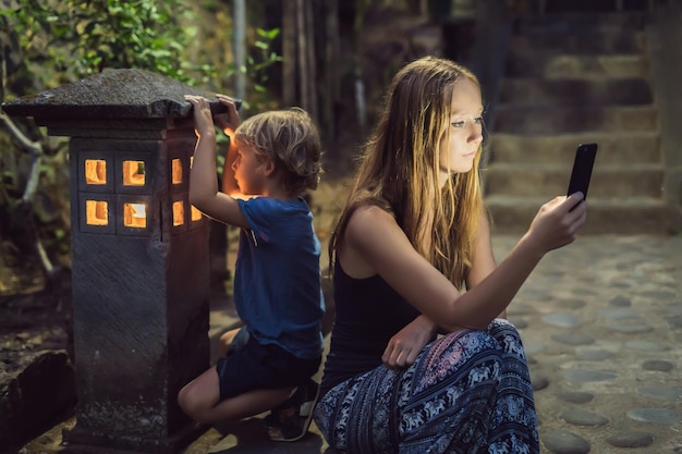 Mom looks into her smartphone, the son looks into the luminous cozy window of the house. The boy lacks Attention, care, love and home comfort