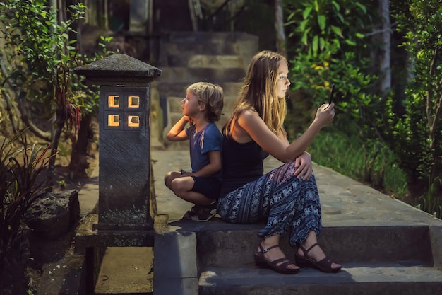 Mom looks into her smartphone, the son looks into the luminous cozy window of the house. The boy lacks Attention, care, love and home comfort