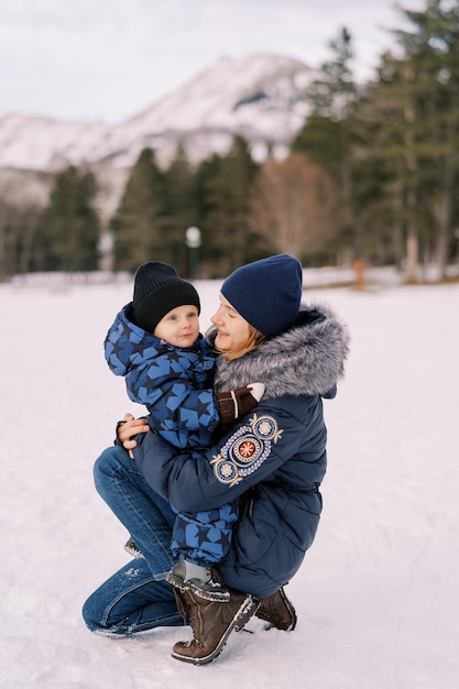 Mom looks at a little boy in her lap while squatting on a snowy lawn