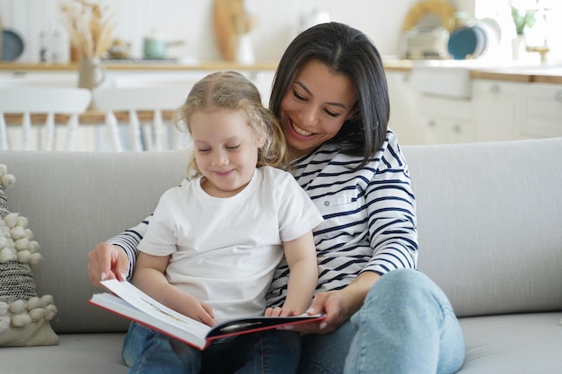 Mom preschool daughter read book together relaxing on cozy sofa enjoying leisure together at home