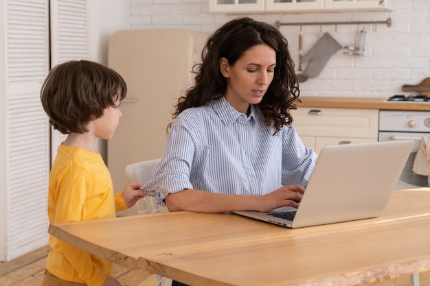 Mom sits by table at home office during lockdown working on laptop and kid distracts and making noise