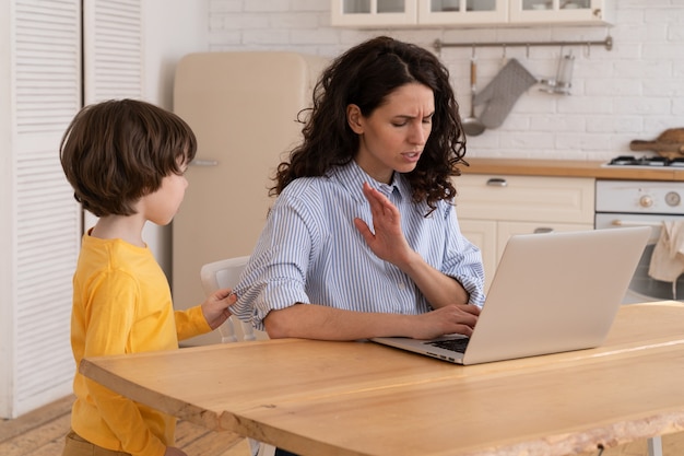 Mom sits by table at home office during lockdown working on laptop. kid distracts and making noise