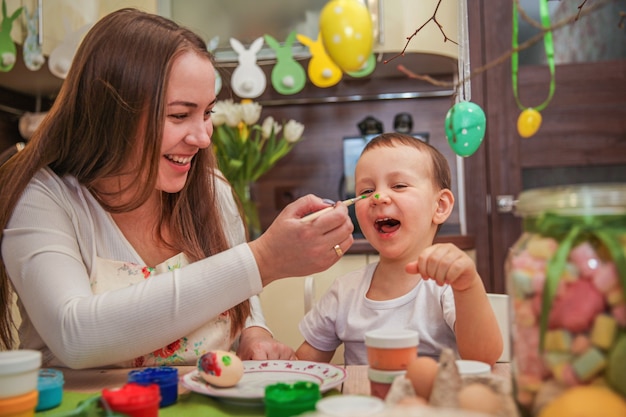 Mom and son in the process of Easter egg painting fool with paints in the decorated kitchen at home