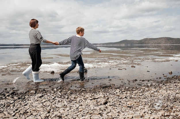 Mom and son on the river bank. Mom and son in vests and rubber boots run merrily on the water. Family intimacy in the open air. Recreation and lifestyle.