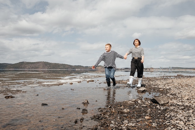 Mom and son on the river bank. Mom and son in vests and rubber boots run merrily on the water. Family intimacy in the open air. Recreation and lifestyle.