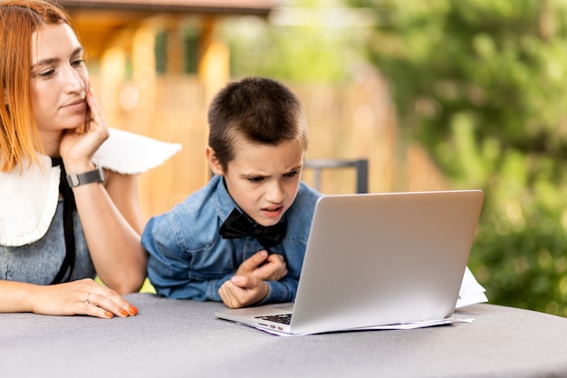 Mom teaches the child at home in the garden, home schooling. Mom helps her son do his homework, stress. Online school lessons via laptop