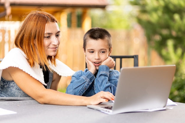 Mom teaches the child at home in the garden, home schooling. Mom helps her son do his homework, stress. Online school lessons via laptop