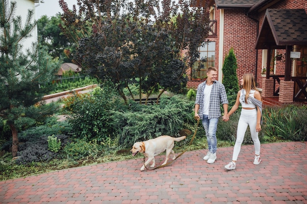 Moment of resting! Young attractive family walking the labrador near the house. Beautiful couple near modern home with dog on the leash.