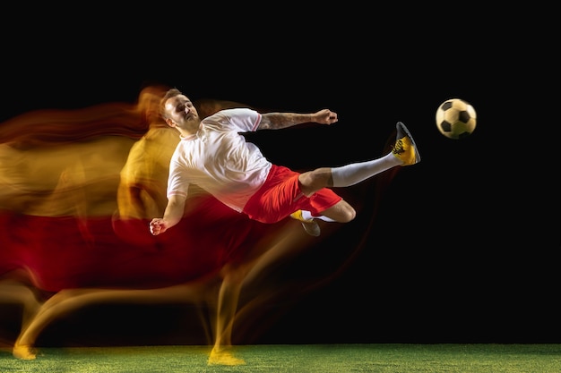 Moment. Young caucasian male football or soccer player in sportwear and boots kicking ball for the goal in mixed light on dark wall. Concept of healthy lifestyle, professional sport, hobby.