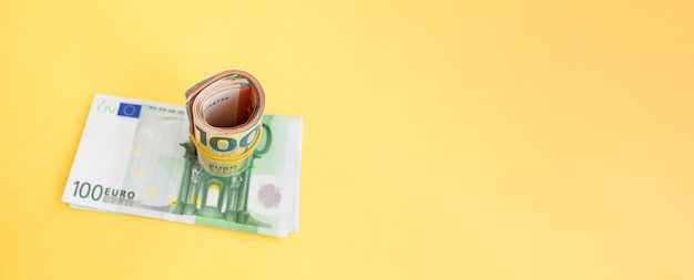 Money roll of euro banknotes with a elastic band on a 100 euro paper bill on a yellow background.