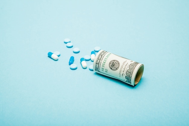 Money rolled up with pills flowing out isolated on blue background, high costs of expensive medication concept. Copy space.