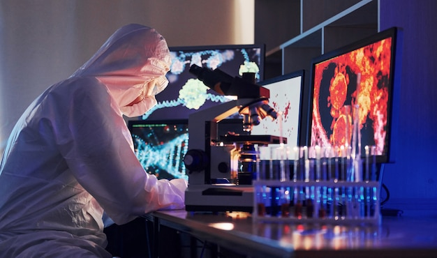 Monitors with information on the table. Scientist in white protective uniform works with coronavirus and blood tubes in laboratory