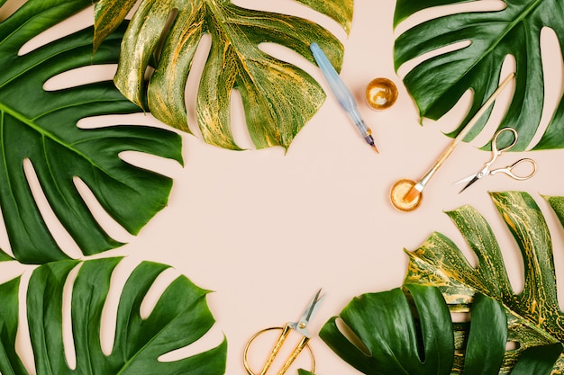 Monstera leaves painted golden with various stationary accessories: brushes, scissors, paint on pastel background