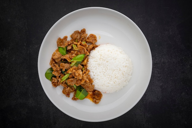 Moo Pad Prik Gaeng, Thai food, stir fried pork with red curry paste, bergamot leaves and yardlong beans with rice in ceramic plate on dark grey texture background