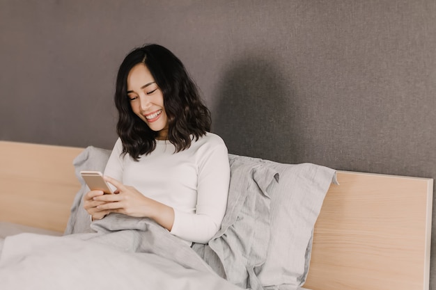 In the morning after wake up, Asian woman is smiling while chatting with her friend in mobile phone on the bed