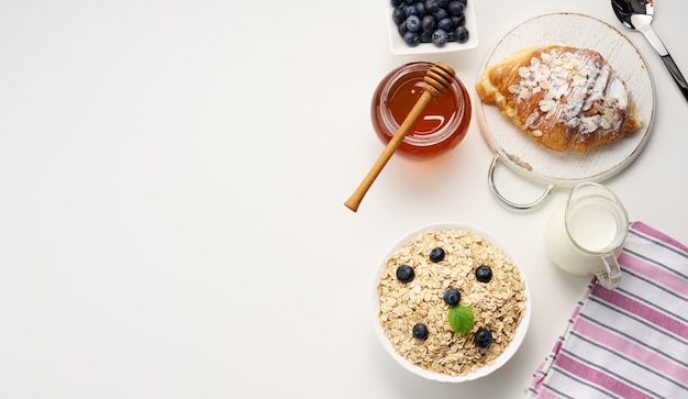 Morning breakfast, raw oatmeal flakes in a ceramic plate, milk in a decanter, blueberries and honey in a jar on a white table, top view, copy space