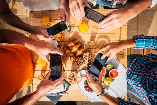 Morning breakfast table full of coffee and food viewed from top vertical above with group of people enjoying and taking pictures with mobile phones together to share on internet. Colorful surface