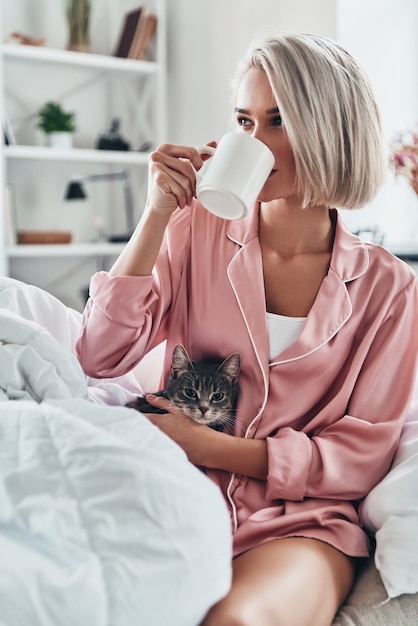 Morning coffee. Attractive young woman drinking coffee and holding her cat while sitting in bed at home