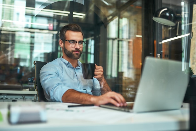 Morning coffee portrait of young and successful bearded man in eyeglasses holding cup of coffee