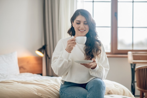 Morning coffee. Pretty young woman sitting on the bed and drinking coffee