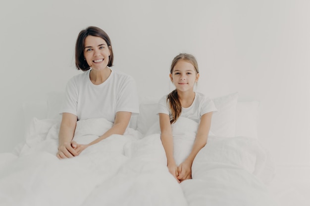 Morning time and awakening concept. Happy smiling mother and daughter sit in comfortable bed, dressed in white t shirts, awake with positive mood, glad to start new day, spend free time in bedroom.