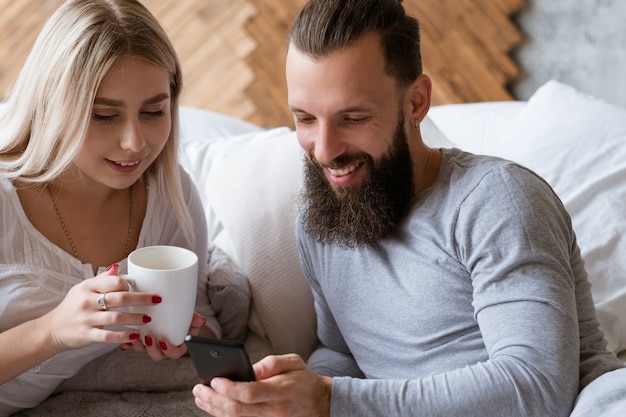 Morning wake up. Young couple ritual. Discussing plans in bed with drink and smartphone.