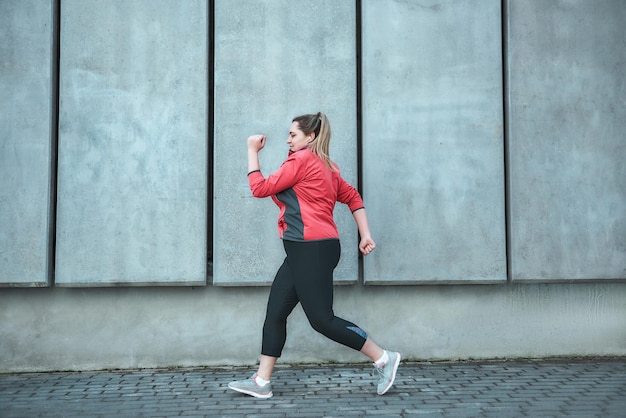 Morning workout. Side view of young plus size woman in sport clothes running outdoors. Healthy lifestyle. Weight losing. Sport concept