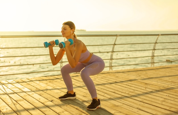 Morning workout. Young slim woman in sportswear practicing squats with dumbbells in hands on the beach at sunrise