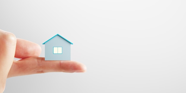 Mortgage concept with small blue house layout on human finger on blank light background with place for advertising poster or your logo mock up