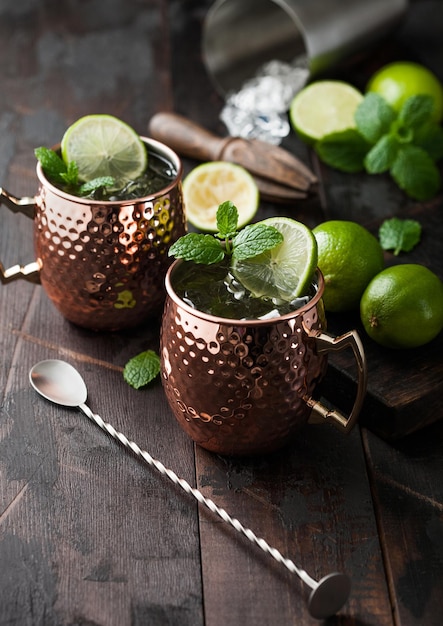 Moscow mule cocktail in a copper mug with lime and mint and wooden squeezer on dark wooden table background and steel shaker and spoon