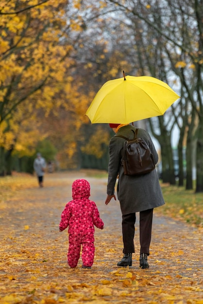 Mother and baby are walking under umbrella along alley of autumn park Back view Vertical frame