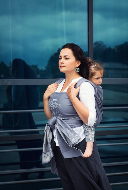 Mother and baby girl sit back in grey wrap sling stay near urban glass with reflection, modern family